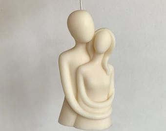 May include: A white candle sculpture of two figures embracing. The figures are stylized and abstract, with simple features. The candle is hanging from a white string.