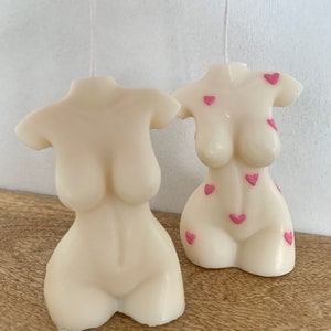 May include: Two white candles shaped like a torso with pink hearts on the surface.