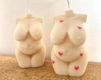 May include: Two nude-colored candles shaped like the human torso with pink hearts on them. The candles are on a wooden surface.