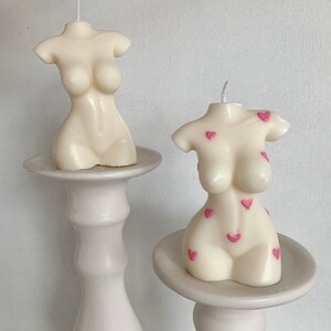 May include: Two white candle sculptures in the shape of a torso with pink hearts on one of the candles. The candles are on white pedestals.
