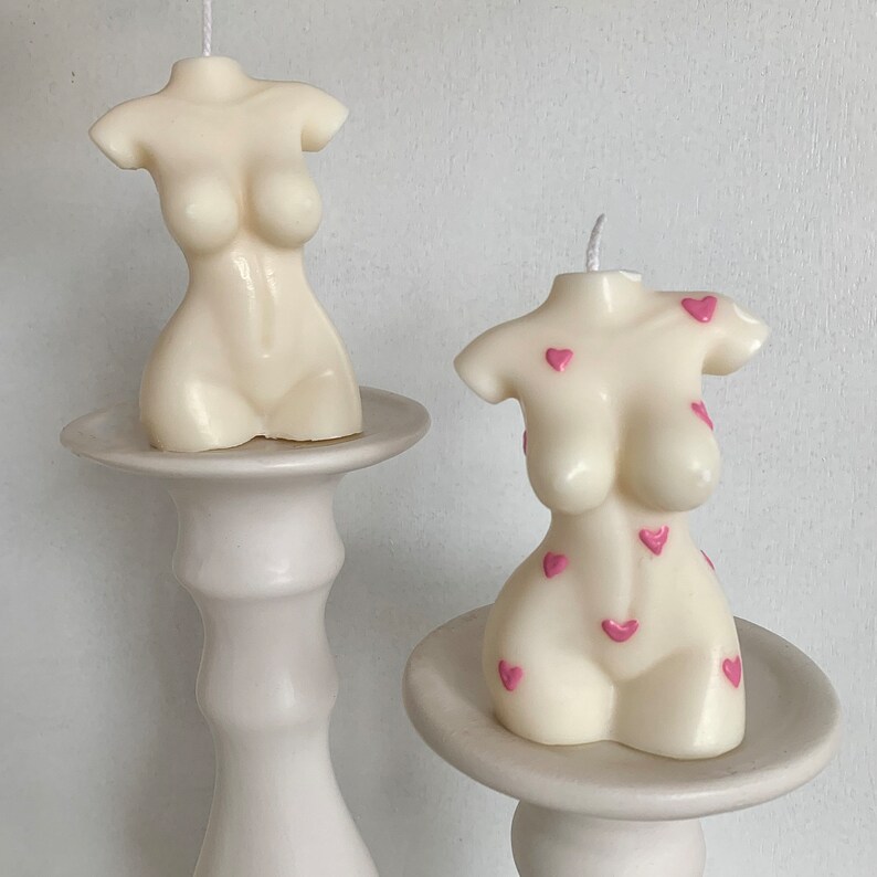 May include: Two white candle sculptures in the shape of a torso with pink hearts on one of the candles. The candles are on white pedestals.