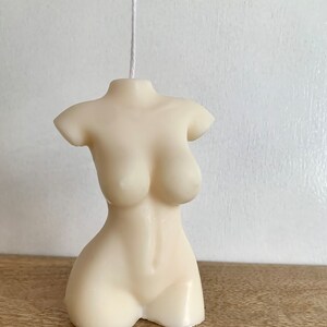May include: A white candle shaped like a human torso. The candle is smooth and has a simple design.