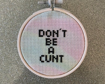 May include: A white embroidery hoop with a pastel tie-dye background and black embroidery that reads "DON'T BE A CUNT".