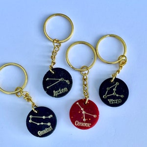 Könnte beinhalten: Vier goldene Schlüsselanhänger mit schwarzen und roten runden Anhängern, die Sternzeichen zeigen. Die Anhänger sind mit "Aries", "Gemini", "Cancer" und "Virgo" beschriftet.