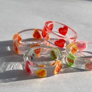 May include: Five clear resin rings with colourful candy hearts embedded in the resin. The hearts have various messages including "Love", "XOXO", "So Cute", "Just Be You", and "I Love You".