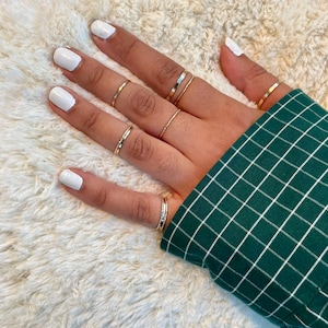 Könnte beinhalten: Eine Hand trägt mehrere dünne goldene Ringe an jedem Finger. Die Hand ruht auf einer weißen, flauschigen Oberfläche. Die Nägel sind weiß lackiert.