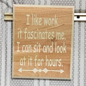 May include: A wooden sign with the text "I like work it fascinates me. I can sit and look at it for hours." The sign has a brown background and white text. There is a small arrow pointing to the right at the bottom of the sign.