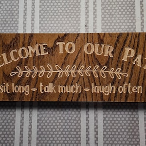 May include: A wooden sign with the text "Welcome to our patio - sit long - talk much - laugh often" in a light brown font. The sign has a dark brown wood grain and a leafy design.