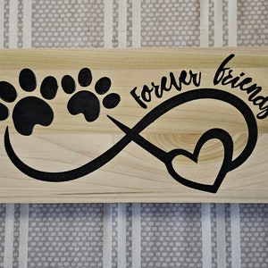 May include: Wooden sign with the words "Forever friends" in black script. The sign features a black paw print design and an infinity symbol with a heart. The sign is made of light-colored wood.