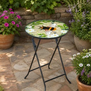 May include: A round mosaic-topped side table with a black metal frame. The tabletop features a bee, white daisies, and green leaves. The table is set on a brick patio with potted plants in the background. The table's diameter is approximately 45 cm.