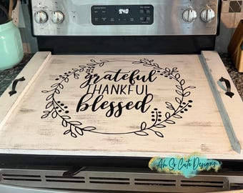 May include: A rectangular white wooden stove cover with a distressed finish, featuring a black circular leaf wreath design and the text "grateful THANKFUL blessed" in the center. It includes two black metal handles on the side edges.
