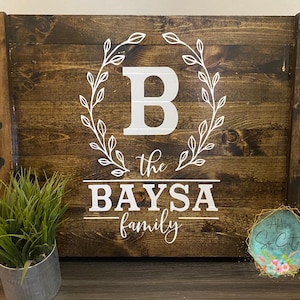 May include: A dark stained wooden serving tray with the text "BAYSAs family" in white. The letter "B" is inside a white floral wreath. The tray has black handles on each side.