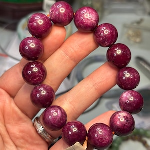 May include: A bracelet of large, round, deep red beads. The beads have a polished, reflective surface and are strung on an orange cord. The bracelet is held in a hand, showing the rich colour and texture of the gemstones.