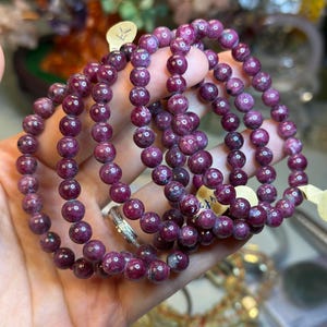 May include: Multiple strands of deep burgundy gemstone bead bracelets. The round beads are approximately 8mm in diameter. The bracelets are held in a hand, highlighting their colour and texture. The background is blurred, suggesting a display setting.