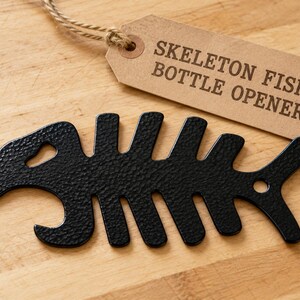 May include: A black metal bottle opener shaped like a fish skeleton with a textured finish. A tan paper tag is attached with twine, featuring the printed text SKELETON FISH BOTTLE OPENER in a dark brown sans-serif font.