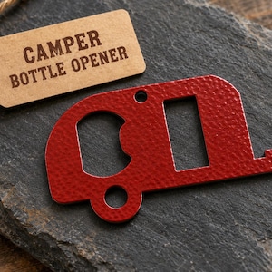 May include: A red metal bottle opener shaped like a vintage camper trailer with a textured finish. A tan paper tag attached with twine features the text CAMPER BOTTLE OPENER printed in dark brown capital letters.