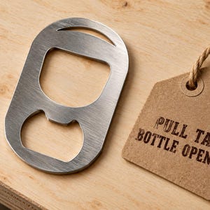Puede incluir: Un abridor de botellas de acero inoxidable cepillado con forma de anilla de lata. Junto al abridor hay una etiqueta de papel marrón claro sujeta con un cordel de yute, con el texto PULL TAB BOTTLE OPENER impreso en tinta negra.