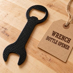 May include: A black, textured metal bottle opener shaped like a wrench, accompanied by a light brown rectangular paper tag attached with a jute string. The tag features the dark brown printed text WRENCH BOTTLE OPENER.