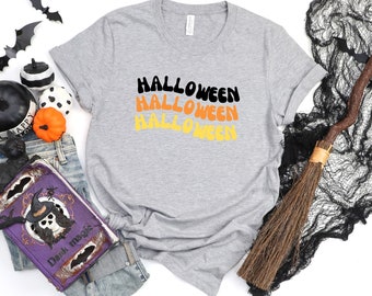 Op de afbeelding: Een grijs T-shirt met het woord "Halloween" drie keer geschreven in een retro-lettertype met oranje, gele en zwarte kleuren.
