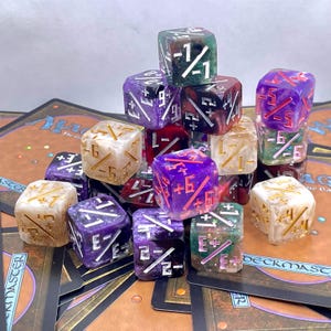 May include: A collection of 12 dice with various colours and patterns. The dice are made of clear resin with embedded glitter and have different numbers and symbols on each side. The dice are arranged in a pyramid shape on a table with playing cards.