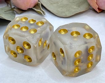 May include: Two translucent dice with gold flecks and gold dots. The dice are on a white surface.