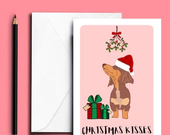 May include: A white greeting card featuring a brown and tan dachshund wearing a red and white Santa hat, standing beside two green and red gift boxes under a sprig of mistletoe, with the text CHRISTMAS KISSES printed in black at the bottom.