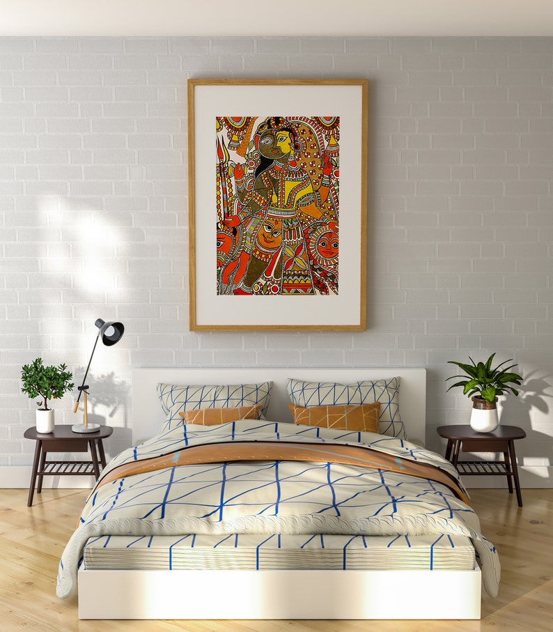 May include: A framed artwork depicting a colorful Indian folk painting with a couple in the center. The painting is set against a white brick wall, above a white bed with a blue and white patterned duvet and a brown throw blanket.