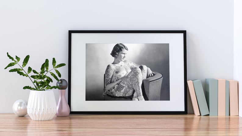 Puede incluir: Fotograf&iacute;a en blanco y negro de una mujer con tatuajes en el cuerpo, sentada en una silla. La mujer mira hacia abajo y hacia la izquierda. La fotograf&iacute;a est&aacute; enmarcada en un marco negro.