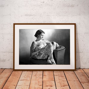 Puede incluir: Fotograf&iacute;a en blanco y negro de una mujer con tatuajes sentada en una silla. La mujer lleva un atuendo sencillo y tiene el pelo corto. La foto est&aacute; enmarcada en un marco marr&oacute;n.