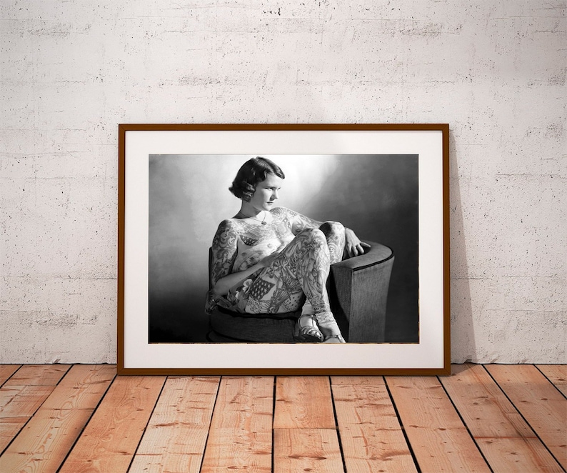 Puede incluir: Fotograf&iacute;a en blanco y negro de una mujer con tatuajes sentada en una silla. La mujer lleva un atuendo sencillo y tiene el pelo corto. La foto est&aacute; enmarcada en un marco marr&oacute;n.
