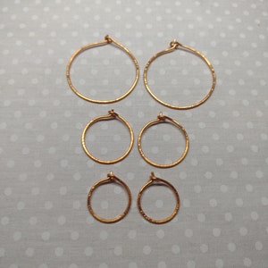 May include: Six pairs of gold-coloured hoop earrings of varying sizes are arranged on a grey and white polka-dotted surface. The earrings have a hammered texture and a simple design, suitable for everyday wear.