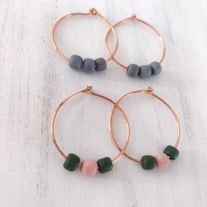 May include: Four pairs of copper hoop earrings. Two pairs have three grey beads, and the other two pairs have a pink bead flanked by two green beads. The earrings are displayed on a white, textured surface.