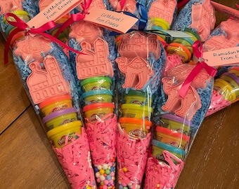 May include: Pink cones filled with colorful sprinkles and Play-Doh containers. The cones are decorated with pink tissue paper and have a tag that says "Eid Mubarak! From: Enaaya".
