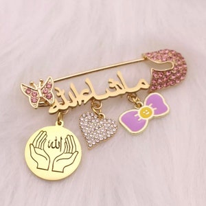 May include: Gold safety pin with a pink butterfly charm, a heart charm with clear rhinestones, a purple bow charm with a yellow smiley face, and a round charm with a gold inscription of hands in prayer with the word "Allah" in Arabic script.