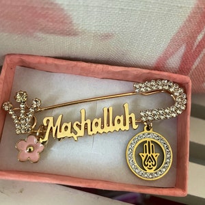 May include: Gold safety pin with a pink flower charm, a charm with the word "Mashallah" and a hamsa hand charm. The charms are decorated with clear rhinestones.