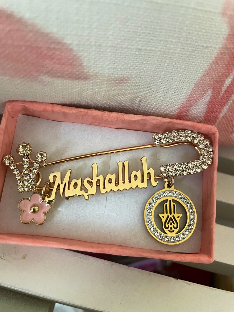 May include: Gold safety pin with a pink flower charm, a charm with the word "Mashallah" and a hamsa hand charm. The charms are decorated with clear rhinestones.