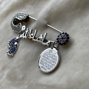 May include: A silver safety pin with a blue and white crystal design. The pin is decorated with the Arabic word "Mashallah" and a small charm with a blue crystal. The pin also has a charm with an oval-shaped pendant with Arabic writing.