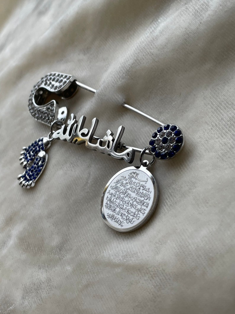 May include: A silver safety pin with a blue and white crystal design. The pin is decorated with the Arabic word "Mashallah" and a small charm with a blue crystal. The pin also has a charm with an oval-shaped pendant with Arabic writing.