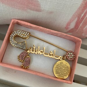May include: A gold safety pin with a pink and white crystal design. The pin has a charm with the Arabic phrase "Masha Allah" and a charm with two pink baby footprints.
