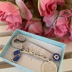 May include: A silver safety pin with blue and clear rhinestones, a charm with the Arabic phrase "Masha Allah" in silver with blue rhinestones, a charm with a blue evil eye with clear rhinestones, and a charm with a round silver disc with Arabic text. The pin is in a light blue box.
