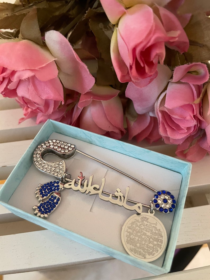 May include: A silver safety pin with blue and clear rhinestones, a charm with the Arabic phrase "Masha Allah" in silver with blue rhinestones, a charm with a blue evil eye with clear rhinestones, and a charm with a round silver disc with Arabic text. The pin is in a light blue box.