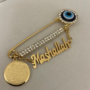 May include: Gold safety pin with a blue evil eye charm, a small gold charm with Arabic script, and a gold charm with the word "Mashallah" in cursive.