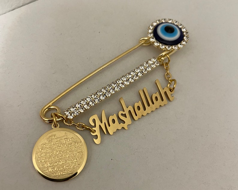 May include: Gold safety pin with a blue evil eye charm, a small gold charm with Arabic script, and a gold charm with the word "Mashallah" in cursive.