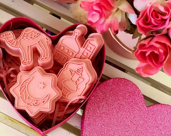 May include: A pink heart-shaped box with a lid, filled with pink cookie cutters in the shapes of a camel, a mosque, a crescent moon and star, and a lantern. The box is sitting on a white wooden surface.