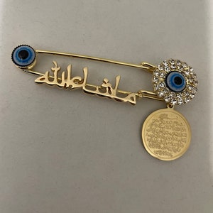 May include: Gold safety pin with a blue evil eye charm, a charm with a blue evil eye surrounded by clear rhinestones, and a round charm with Arabic script.