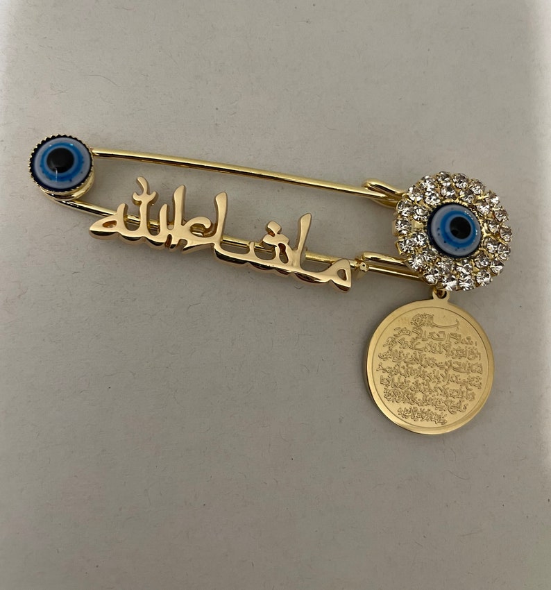 May include: Gold safety pin with a blue evil eye charm, a charm with a blue evil eye surrounded by clear rhinestones, and a round charm with Arabic script.