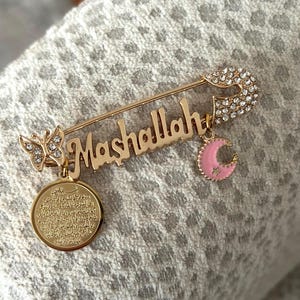 May include: Gold safety pin with a charm that says "Mashallah" in Arabic script. The pin is decorated with clear rhinestones and has a pink crescent moon charm and a butterfly charm with clear rhinestones.