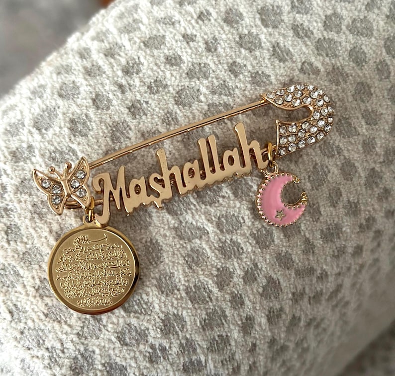 May include: Gold safety pin with a charm that says "Mashallah" in Arabic script. The pin is decorated with clear rhinestones and has a pink crescent moon charm and a butterfly charm with clear rhinestones.