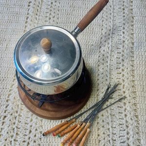 May include: A vintage fondue set with a stainless steel pot, wooden handle, and lid. It sits on a wooden base with a black metal stand. Includes a set of fondue forks with wooden handles and colored tips. The background is a cream-colored crocheted blanket.