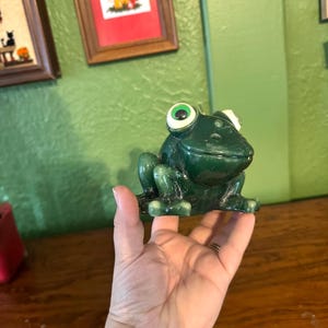May include: A green frog figurine with large, white-rimmed eyes. The frog is sitting upright, with its front legs extended. The figurine is made of a glossy material and is a decorative item.